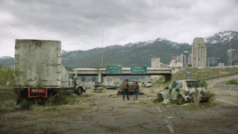 Post-apocalyptic highway scene from HBO's The Last of Us with overgrown vehicles, abandoned city, and mountains in the background.