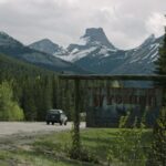 A vehicle drives into Wyoming through a forested highway, with dramatic mountain peaks hinting at The Last of Us journey ahead.