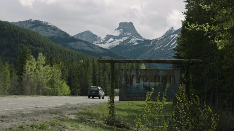 A vehicle drives into Wyoming through a forested highway, with dramatic mountain peaks hinting at The Last of Us journey ahead.